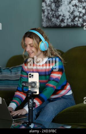 Smiling young woman in vibrant sweater wears headphones while using a laptop, indicative of remote smart working at home Stock Photo