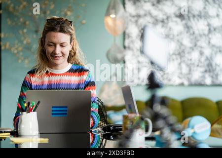 A smiling young woman engages in smart working from an eclectically decorated home interior, embracing a comfortable and creative work environment Stock Photo