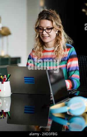 A joyful young woman in glasses works from home on her laptop, embodying modern smart working trends and enjoying the comfort of remote work, with a c Stock Photo