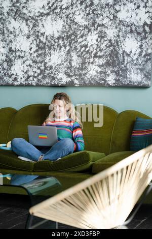 A content woman works remotely from home, sitting on a vibrant green sofa with a colorful sweater and laptop Stock Photo