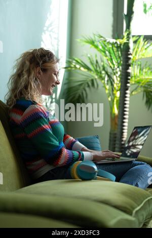 A cheerful young woman enjoys smart working from a cozy home environment, seated on a green armchair with laptop, surrounded by lush houseplants Stock Photo