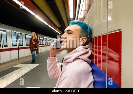 A young male with vibrant blue hair and wearing a pink hoodie is engaged in a conversation on his mobile phone while waiting at a subway station Stock Photo