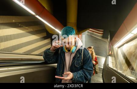 A stylish young man with striking blue hair stands on an escalator, wearing sunglasses and adjusting them while looking at his smartphone Stock Photo
