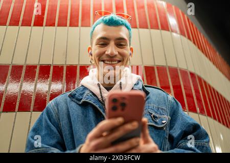 A cheerful young man with vibrant blue hair smiles while using his smartphone, standing against a red tiled subway station wall Stock Photo