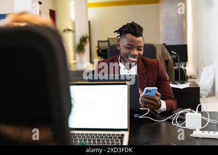 A young man in a stylish burgundy blazer smiles while using his smartphone and laptop at a modern, well-lit office workspace Stock Photo