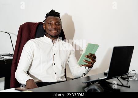 A cheerful young businessman in a white shirt sits at his office desk, smiling as he checks his smartphone, with a laptop and workstation visible in t Stock Photo