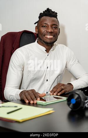 A cheerful young man in a white shirt sits at his office desk, smiling warmly while engaging in work-related activities He is surrounded by office ess Stock Photo