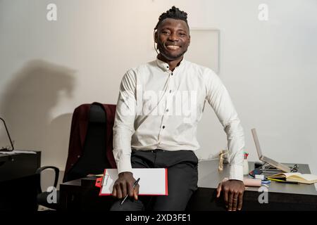 A young man in a white shirt, positioned confidently at his desk with a red folder, exhibits a radiant smile, displaying professionalism and approacha Stock Photo