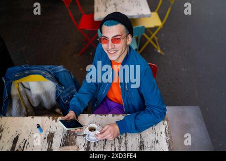 A young man with blue hair and red sunglasses smiles while using his phone and drinking coffee at a street cafe, seated at a weathered white table sur Stock Photo