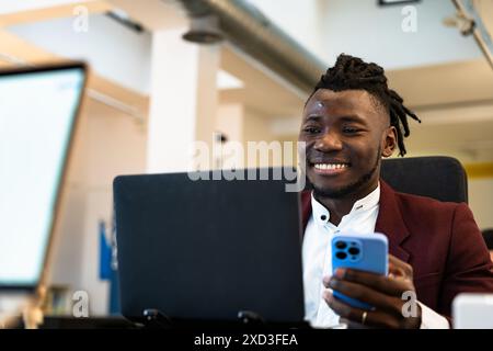 A cheerful young Black man in a stylish red blazer uses a smartphone and laptop simultaneously at a modern office setting, symbolizing multitasking an Stock Photo