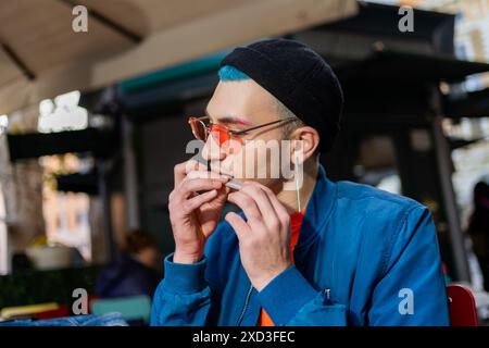 A stylish young man enjoys a hamburger at a street cafe He’s dressed in a modern blue jacket and sports teal hair under a black beanie, with orange-ti Stock Photo