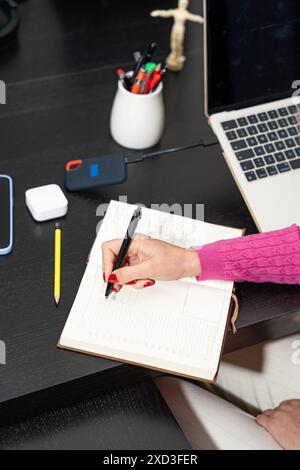 A close-up of a woman's hand using a pencil to write in a planner while seated at a modern desk, surrounded by various gadgets and a laptop Stock Photo