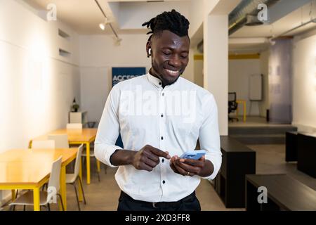 A young, cheerful man dressed in a white shirt stands in a well-lit office space, happily using his smartphone Stock Photo