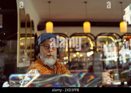 A fashionable senior man in a colorful shirt and cap working on his laptop at a café, headphones on, embodying modern remote work lifestyle Stock Photo