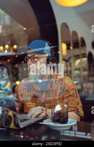 A modern senior with a beard works remotely, using headphones and a laptop at a coffee shop, exemplifying an active lifestyle and nature illustrations Stock Photo