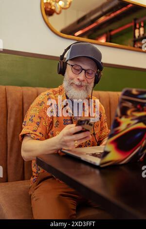 A modern, stylish senior man enjoys remote work as he listens to music through headphones, smiling while using his smartphone and laptop in a cozy caf Stock Photo