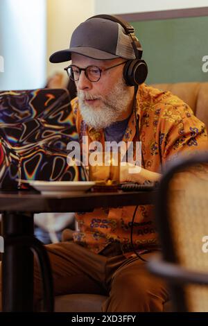 A contemporary senior male engages in remote work, showing modern retirement lifestyle Wearing headphones, he's focused on a laptop at home Stock Photo