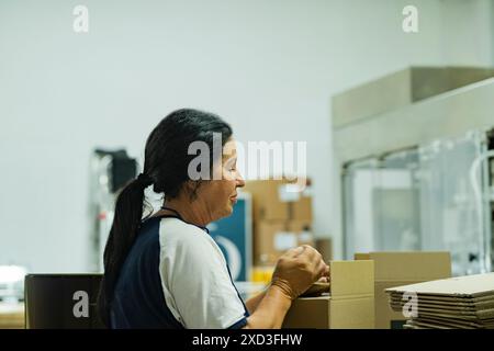 A mature female worker with a braid is focused on packaging items in a distribution warehouse, illustrating traditional labor in industrial settings Stock Photo