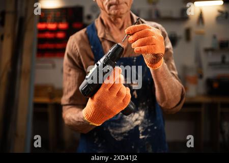 Elderly man inserting a drill bit into an electric drill in his workshop. He is wearing brown shirt, blue apron, and orange gloves, preparing for Stock Photo