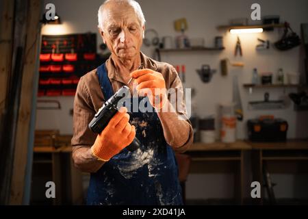 Elderly man inserting a drill bit into an electric drill in his workshop. He is wearing brown shirt, blue apron, and orange gloves, preparing for Stock Photo
