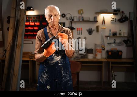 Elderly man inserting a drill bit into an electric drill in his workshop. He is wearing a brown shirt, blue apron, and orange gloves, preparing for a Stock Photo