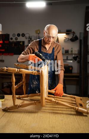 Elderly man in a workshop using a drill with a special attachment to remove old varnish from a wooden chair. He is wearing protective goggles and Stock Photo