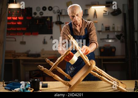 An elderly man in orange gloves and an apron repairs an old wooden chair in a workshop. He wraps the legs of the chair with tape before painting. Stock Photo