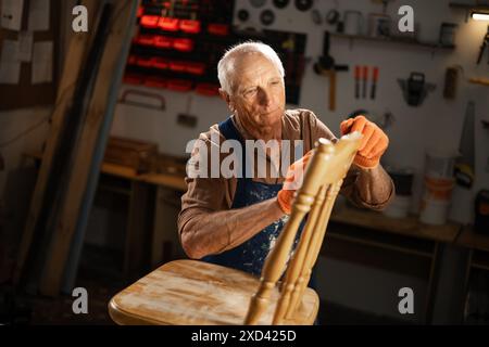 An elderly man in orange gloves and a blue apron sands an old wooden chair in his workshop. He is in his workshop and is focused on preparing the Stock Photo