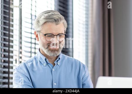 Smiling mature businessman wearing glasses and blue shirt, working on laptop with white blinds in the background. Stock Photo
