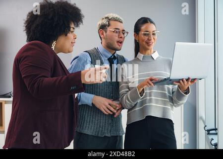 Three people are standing with a laptop watching a funny video in the office,There are two women and a man, one of them holds the laptop showing it to Stock Photo