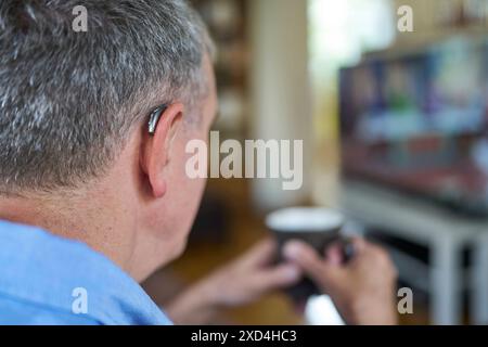 Close Up Of Mature Man Wearing Wireless Hearing Aid Watching TV At Home  Relaxing With Hot Drink Stock Photo
