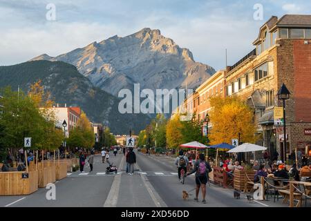 Banff, Alberta, Canada - September 28 2020 : Tourists are walking and eating outside on Banff Avenue in autumn evening. Cascade Mountain over rosy sky Stock Photo