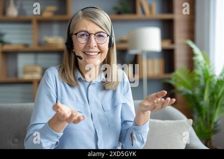 Senior woman wearing a headset during a video call, smiling and talking happily, sitting in a cozy living room. Stock Photo