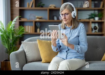 Senior woman sitting on a cozy couch in the living room using a tablet with headphones, enjoying technology and staying connected. Stock Photo