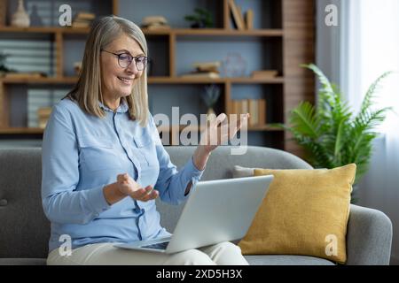 Senior woman talking and gesturing during a video call, using a laptop, sitting on a sofa in a cozy living room Stock Photo