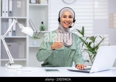 Smiling customer service representative wearing a headset gives a thumbs up while working at her office desk with a laptop ,and lamp in a modern workspace. Stock Photo