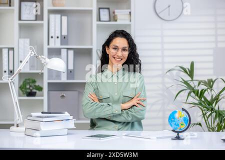 Confident female teacher smiling while standing in a modern classroom with school supplies and books, representing education and learning. Stock Photo