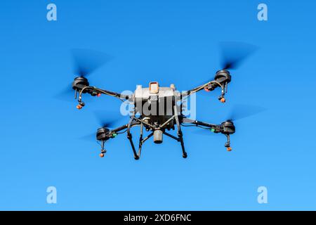 A pesticide sprayer drone flying in the air, against a clear blue sky. The drone has four propellers, a spray tank, and is equipped with nozzles for precise pesticide application. Stock Photo