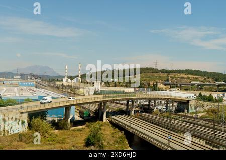 An expansive view showcasing a highway overpass and parallel rail tracks cutting through an industrial zone with factories under a clear sky. Stock Photo