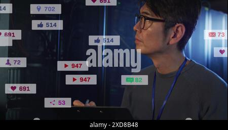 Image of social media icons over asian male engineer writing on clipboard at server room Stock Photo