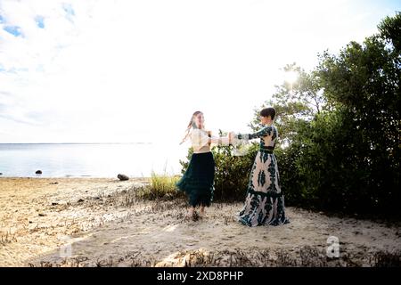 Two women in elegant dresses dancing together on a sandy beach n Stock Photo