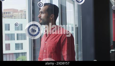 Image of bar graph icons over thoughtful middle eastern man looking out of window at office Stock Photo