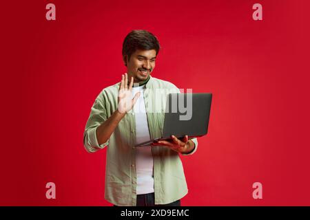 Young, confident Indian man dressed casual outfit holds meeting online using laptop against red studio background. Stock Photo