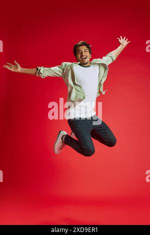 Dynamic photo of young Indian man jumping in mid-air of joy and happiness against red studio background. Positive emotions. Stock Photo