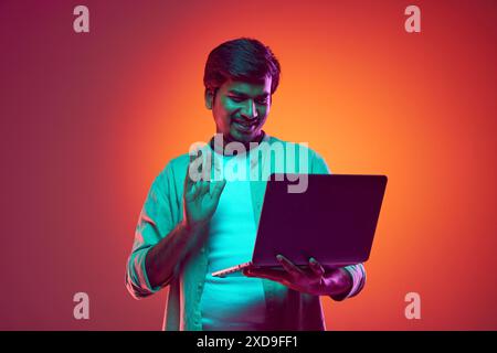 Young, confident Indian man dressed casual outfit holds meeting online using laptop in neon light against gradient studio background. Stock Photo