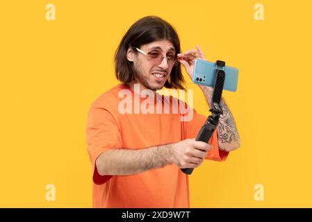 Cheerful middle eastern guy taking photo by smartphone with selfie stick on yellow background. Stock Photo