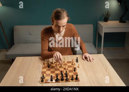 Young man with focused expression playing chess at home, sitting at a wooden table in a relaxed indoor setting. atmosphere as he contemplates his next Stock Photo