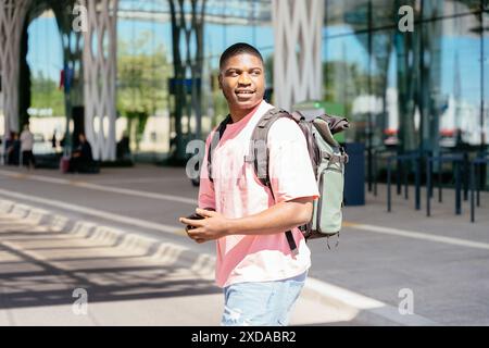 A man wearing a pink shirt and a backpack is walking down a sidewalk. He is holding a cell phone in his hand. Stock Photo
