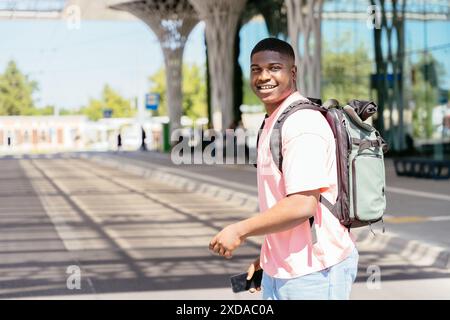 African man wearing a pink shirt and a green backpack is smiling and holding a cell phone. He is walking down the street, possibly heading to a Stock Photo