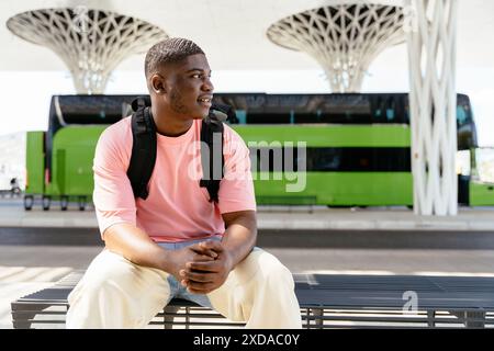 A man in a pink shirt sits on a bench next to a green bus. He is wearing a backpack and he is waiting for the bus Stock Photo